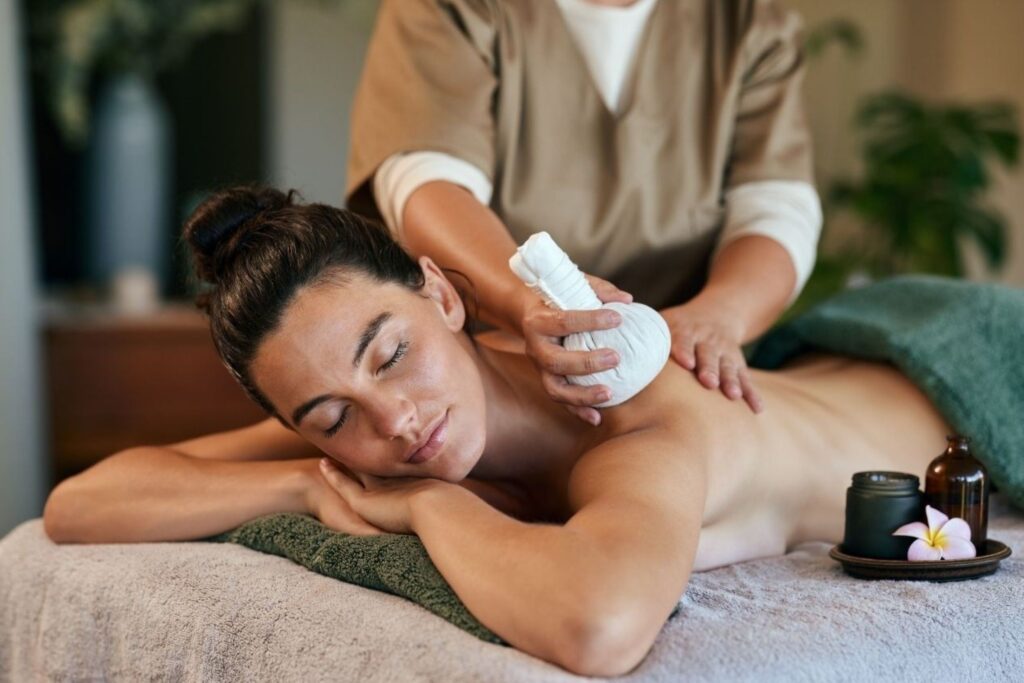 Relaxed woman lying face down on a massage table while a therapist wraps her forearm with a cloth wrap in a spa setting.