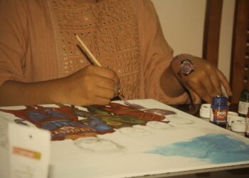 A woman carefully creating a Madhubani painting on thick white paper with a fine-tipped brush
