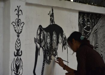A woman in Maharashtra creating Warli art on brown paper using traditional white paint and a fine brush