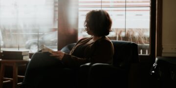 A woman sits in a cozy reading nook near a rain-streaked window, wrapped in a cotton throw, holding a book