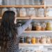 indian-woman-arranging-spice-jars-unsplash - INDIAwebzine A woman carefully organising her home spice pantry with colourful Indian regional spices on a wooden shelf