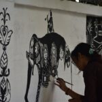 Una mujer en Maharashtra creando arte Warli en papel marrón usando pintura blanca tradicional y un pincel fino
